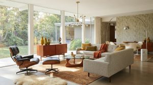 Na obrazie przedstawiony jest nowoczesny salon urządzony w stylu mid-century modern, z dużymi przeszkleniami, przez które widać zielony ogród